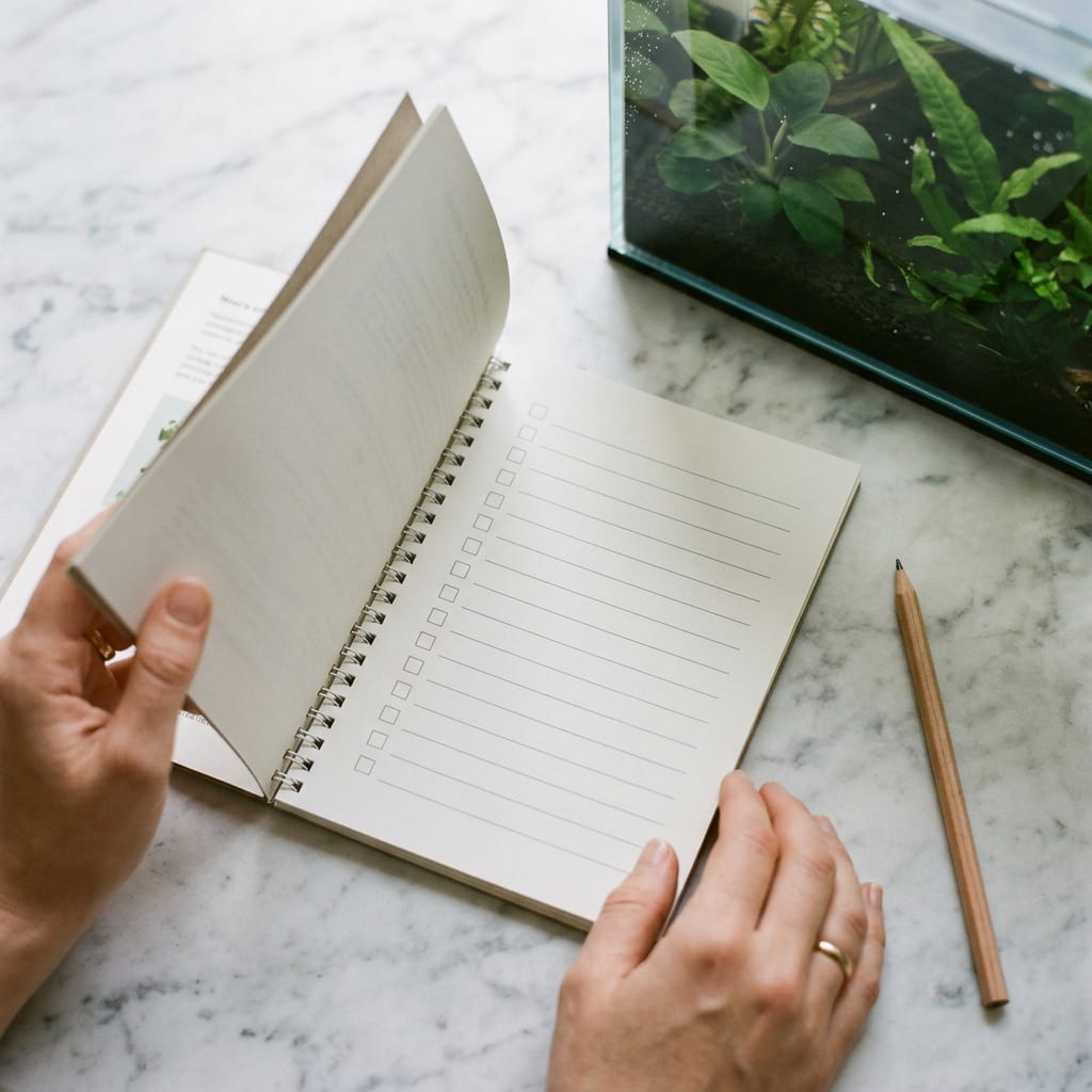 Illustrative image: hands turning a page of a printed beginner guide with a checklist, next to a planted freshwater tank.